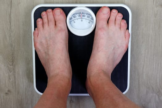 Old weight scale stood on my adult male seeing point of view of scale standing on. Adult white male weighing his self on an old worn black and white weight scale on a wood grain design vinyl floor.