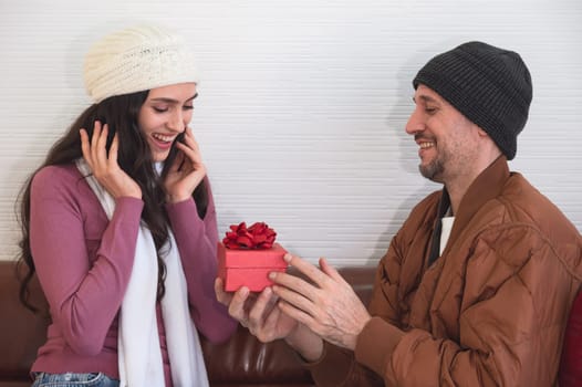 Man surprising his girlfriend with red gift box on couch in living room. Couple celebrating holidays, anniversary or birthday together at home.