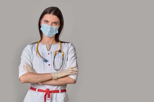 A young female doctor in a white coat with a stethoscope and a medical mask on a gray isolated background. copy space. High quality photo