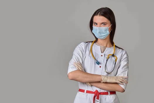 A young female doctor in a white coat with a stethoscope and a medical mask on a gray isolated background. copy space. High quality photo