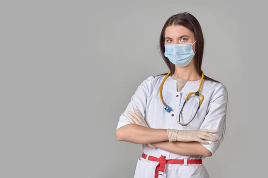 A young female doctor in a white coat with a stethoscope and a medical mask on a gray isolated background. copy space. High quality photo