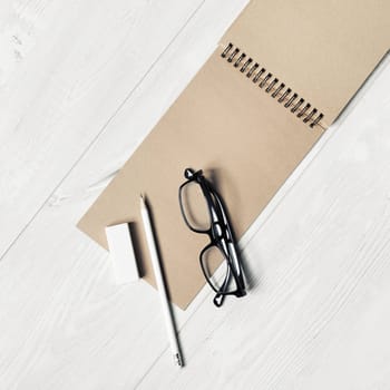 Blank kraft notebook, glasses, pencil and eraser on light wooden background. Template for placing your design. Top view. Flat lay.