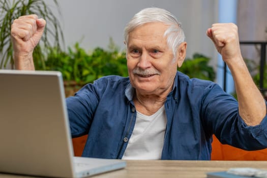 Happy senior old man at home couch with laptop scream in delight raise hands in triumph winner gesture celebrate success win money in lottery. Excited elderly mature grandfather get online good news
