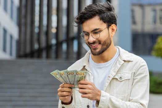 Happy young Indian man counting money cash holding dollars banknotes, bills budget count salary profit reward bribe outdoors. Successful rich Arabian Hindu guy in downtown city street. Business people