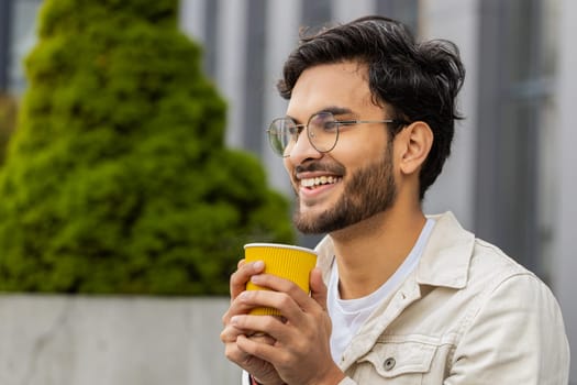 Happy young Indian man enjoying morning coffee hot drink and smiling outdoors. Relaxing, taking a break. Arabian Hindu guy in urban city downtown street, drinking coffee to go. Town lifestyles outside