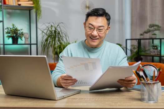 Happy Asian man freelancer work with documents at home analyzing information read business papers prepare financial report. Guy winner shocked by victory, good success news sits on couch at table desk