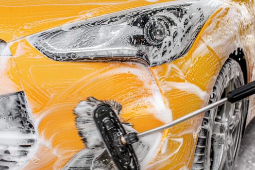 Front headlight and bumper of yellow car being washed in self service carwash, brush leaving strokes in shampoo and foam.