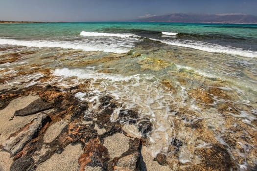 Sand on large rocks, crystal clear turquoise sea with white waves and land in background. Chrissi , Greece