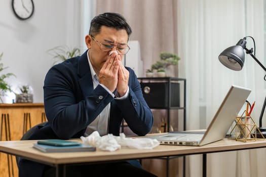 Sick ill Asian middle-aged businessman in suit suffers from cold flu fever or allergy working on laptop at home office. Overworked freelancer employee mature man blows sneezes wipes snot into napkin