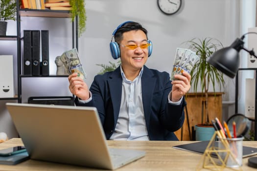 Asian Chinese businessman holding fan of cash money dollar banknotes celebrate, dance, success career, lottery jackpot game winner, big income, wealth at office workplace desk. Remote distant working