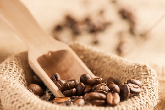 Close-up view of roasted coffee beans on burlap sack with small wooden scoop in front of blurred background.
