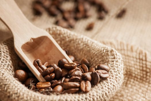 Close-up view of roasted coffee beans on burlap sack with small wooden scoop in front of blurred background.