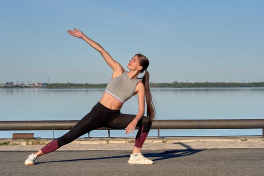 A young woman does stretching on the pier in the early morning, against the background of the river. High quality photo