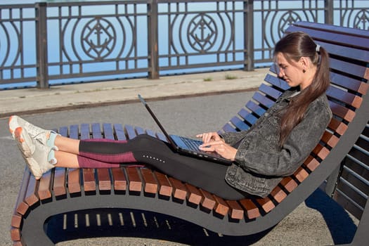 Young woman sitting on a bench with a laptop and working on a sunny summer day. High quality photo