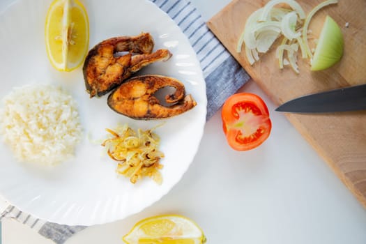 Fried fish with onions and rice is on a plate, on a waffle towel, next to it is a cutting board with chopped onions, lemon and tomato on a white background. High quality photo