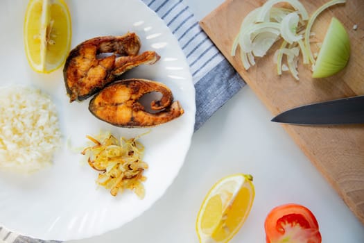 Fried fish with onions and rice is on a plate, on a waffle towel, next to it is a cutting board with chopped onions, lemon and tomato on a white background. High quality photo
