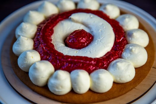 The baked cake cake lies on a wooden substrate and on a white stand, the cake is decorated with white cream and raspberry jam, in a circle, circles alternate on a dark background. High quality photo