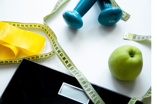 An apple, a scale, a fitness elastic band and dumbbells lie on a white background, they are entwined with a measuring tape. High quality photo