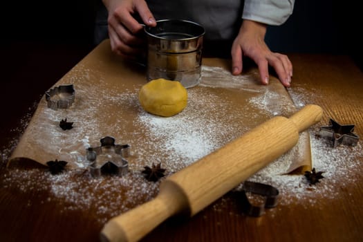 A table with baking parchment lying on it, scattered flour, rolling pin, dough and baking molds in dark colors. The cook's hands are visible in an apron, which is holding a sifter with flour. High quality photo