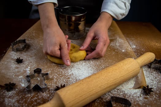 In the foreground is a table with baking parchment lying on it, on which there is dough that the cook's hands are crumpling, a rolling pin, baking molds and flour are scattered around. High quality photo