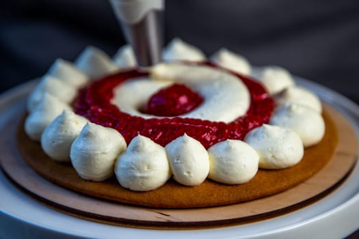The baked cake cake lies on a wooden substrate and on a white stand, the cake is decorated with white cream and raspberry jam, in a circle, circles alternate, a pastry bag with cream on a dark background is placed above the cream cake. High quality photo
