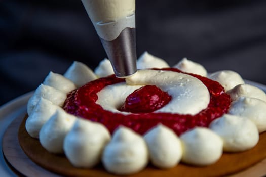 The baked cake cake lies on a wooden substrate and on a white stand, the cake is decorated with white cream and raspberry jam, in a circle, circles alternate, a pastry bag with cream on a dark background is placed above the cream cake. High quality photo