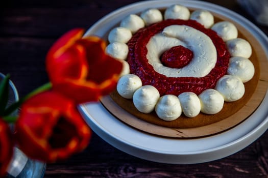 The baked cake cake lies on a wooden substrate and on a white stand, the cake is decorated with white cream and raspberry jam, in a circle, circles alternate, a bouquet of red tulips on a dark background is visible nearby. High quality photo