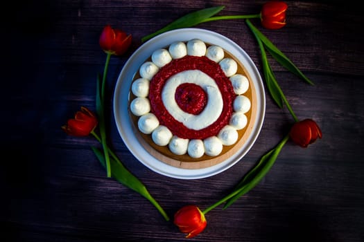 Honey sponge cake with a decoration of white air cream and layers of red raspberries stands on a white platter on a dark wooden background, tulips lie around in a circle. High quality photo