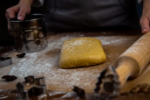 A rectangle of dough lies on baking parchment, a rolling pin and cookie cutters are lying next to it, everything is strewn with flour, a cook is standing in the background and holding a rolling pin and flour powder. High quality photo