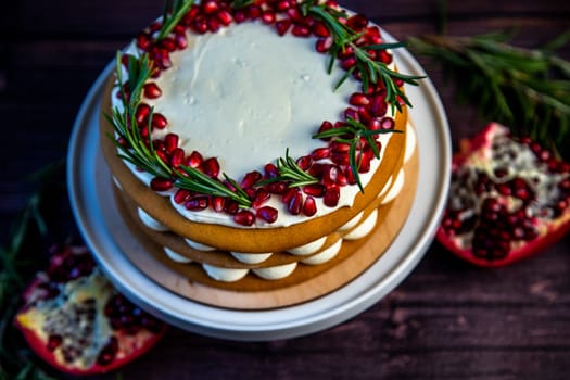 Cake with white cream and honey brown cakes lies on a white stand, decorated with pomegranate and rosemary, against a dark tree. High quality photo
