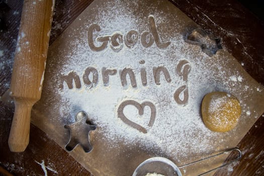 On the baking paper on the flour is written Good morning and heart, against the background of dough, rolling pins and cookie cutters. High quality photo