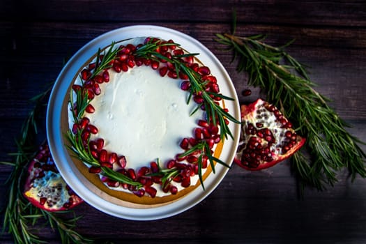 Cake with white cream and honey brown cakes lies on a white stand, decorated with pomegranate and rosemary, against a dark tree. High quality photo