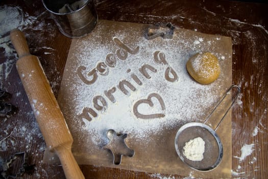 On the baking paper on the flour is written Good morning and heart, against the background of dough, rolling pins and cookie cutters. High quality photo