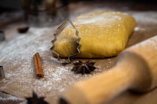 A cookie cutter in the form of a Christmas tree is leaning on the dough, cinnamon and cloves are lying next to it, flour, other cookie cutters and a rolling pin for dough are visible in the background, and everything is in flour. High quality photo