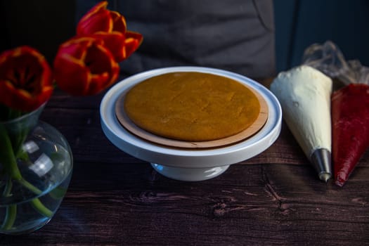 The baked cake cake lies on a white stand, pastry bags with cream and jam are next to it, a bouquet of flowers on a dark background is visible from the side in a vase. High quality photo
