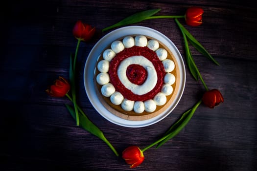 Honey sponge cake with a decoration of white air cream and layers of red raspberries stands on a white platter on a dark wooden background, tulips lie around in a circle. High quality photo