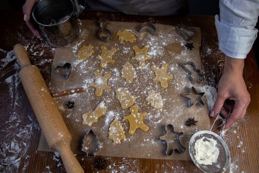 There are ready-made cookies of various shapes on the baking parchment, a rolling pin, cookie cutters and a sieve with flour are lying nearby, flour is scattered everywhere, the hands of the pastry chef are visible. High quality photo