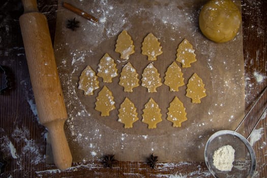 A lot of cookies cut out of the dough in the form of Christmas trees lies on the baking paper in flour, there is a rolling pin and dough next to it. High quality photo