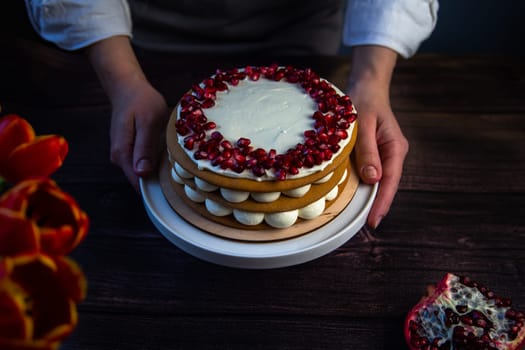 The cake stands on a white stand, decorated with cream and pomegranate on top, it is held by the hands of a pastry chef, there is a pomegranate next to it and there are flowers on a dark background. High quality photo