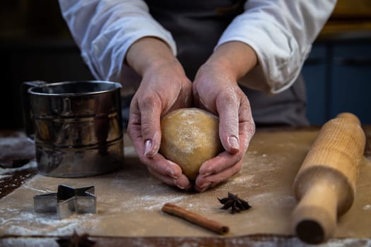 In the hands of a pastry chef, a round piece of dough, all in flour, a rolling pin, a mold with flour, cloves and cinnamon, all in dark colors, lies next to it. High quality photo