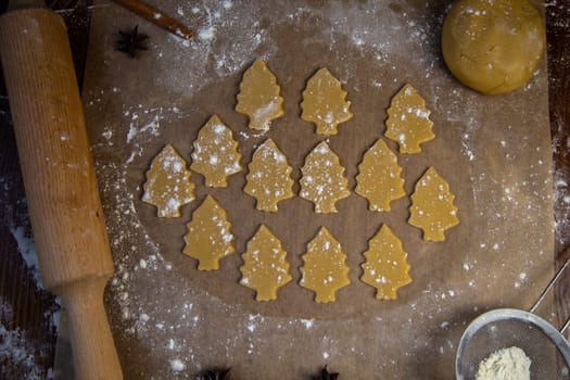 A lot of cookies cut out of the dough in the form of Christmas trees lies on the baking paper in flour, there is a rolling pin and dough next to it. High quality photo