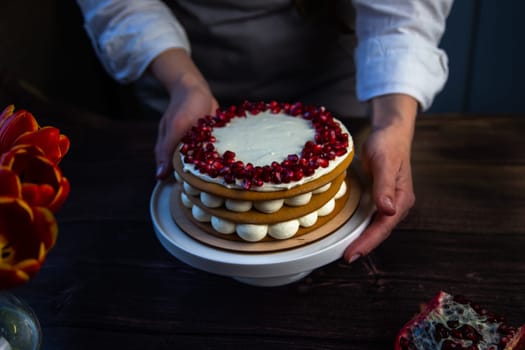 The cake stands on a white stand, decorated with cream and pomegranate on top, it is held by the hands of a pastry chef, there is a pomegranate next to it and there are flowers on a dark background. High quality photo