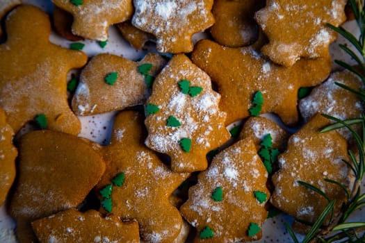 Cookies are on a plate of different shapes, in the foreground there is a cookie in the form of a Christmas tree, decorated with powdered sugar like snow and small green Christmas trees against the background of the rest of the cookies, a branch of rosemary is visible from the side. High quality photo