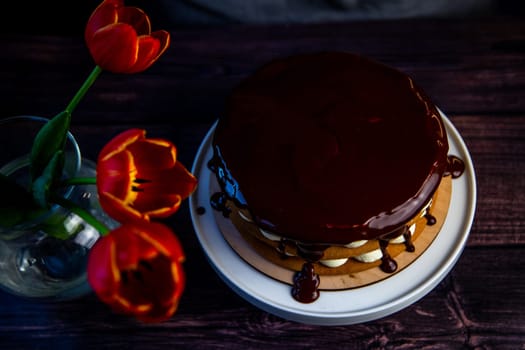 Red tulips stand in a glass vase near the cake, poured on top with chocolate icing on a dark background. Top view. High quality photo