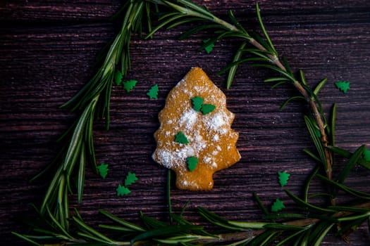 Cookies in the form of a Christmas tree, sprinkled with powdered sugar and small Christmas trees on top, lies on a dark wooden background surrounded by grained rosemary. High quality photo