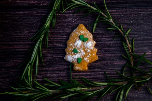 Cookies in the form of a Christmas tree, sprinkled with powdered sugar and small Christmas trees on top, lies on a dark wooden background surrounded by grained rosemary. High quality photo