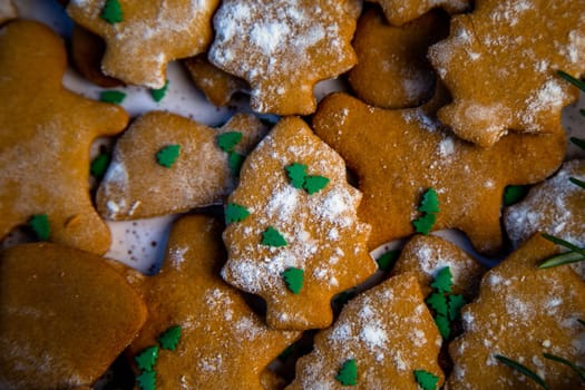 Cookies are on a plate of different shapes, in the foreground there is a cookie in the form of a Christmas tree, decorated with powdered sugar like snow and small green Christmas trees against the background of the rest of the cookies, a branch of rosemary is visible from the side. High quality photo