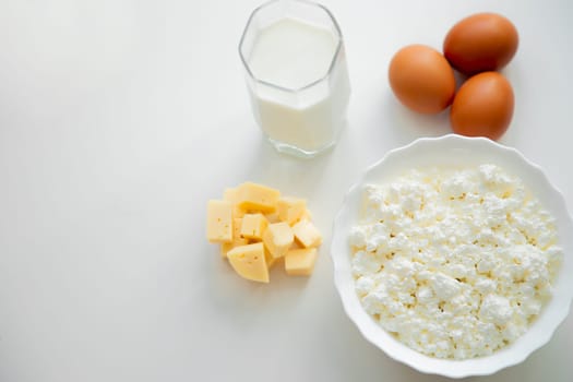 A white bowl with cottage cheese, eggs, diced cheese and a glass of milk stand on a white background. . High quality photo