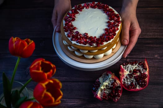 The cake stands on a white stand, decorated with cream and pomegranate on top, it is held by the hands of a pastry chef, there is a pomegranate next to it and there are flowers on a dark background. High quality photo