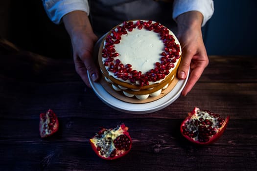 A cake with sponge cakes, decorated with white cream and pomegranate seeds on top, is held in a circle by a pastry chef against a background of dark wood and pomegranate. High quality photo